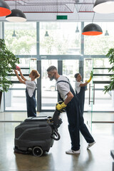 Wall Mural - side view of man with floor scrubber machine near multicultural women cleaning office hall.