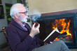 © Solid photos - Senior man smoking pipe and reading book by fireplace in winter
