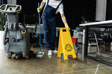 Wall Mural - cropped view of man with mop holding caution sign board near floor scrubber machine.