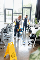 Wall Mural - happy woman washing floor with mop near caution board and mixed race colleague.