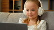 © Yuliia - Funny little blonde girl child cute schoolgirl pretty beautiful kid young blogger wears headphones sit on sofa talking to webcam making online video call recording vlog having fun waving hand greeting