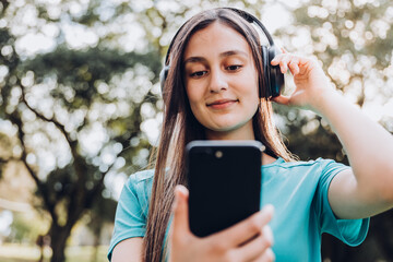 Wall Mural - Teenage girl wearing turquoise t shirt, using headphones and setting a music playlist on her smartphone in the park