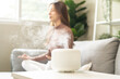 © KMPZZZ - Air humidifier, calm blurred woman, girl sitting on couch lotus pose put hands practice meditation do yoga exercise at home. Aromatherapy steam scent from essential oil diffuser in living room at home