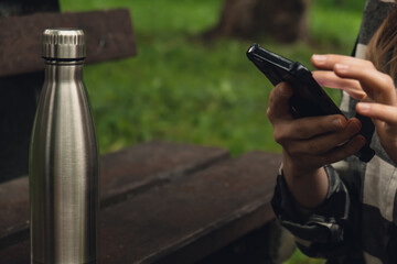 Mobile phone social media. Reusable steel thermo water bottle on wooden bench. Sustainable lifestyle. Plastic free zero waste free living. Go green Woman student work study with phone in park