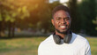 © lenblr - Smiling African American man wearing black headphones walks in green park. Black adult enjoys walking exploring great destination at sunlight