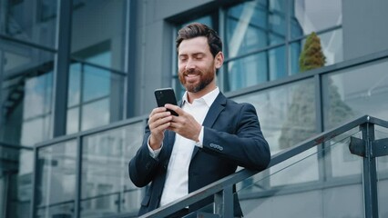 Smiling man standing outdoors holding mobile phone browsing social network happy pensive businessman thinks dreams writes email message on telephone chatting with client or friend using smartphone