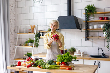 Wall Mural - beautiful elderly gray haired senior woman cook in cozy kitchen with fresh organic vegetables, tomatoes, cabbage, lettuce, cucumbers on table cooking healthy vegetable salad, healthy food active life