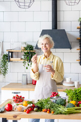 Wall Mural - beautiful elderly grey haired senior woman in cozy kitchen takes vitamin supplement in capsule, drinks pure water from glass, biologically active food additives for full life, healthy food active life