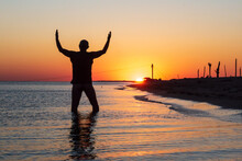 Silhouette Of Man At Sunset In Sea Water With Hands Raised Towards Setting Sun. Worship, Praise, Thanksgiving, Freedom And Love To Nature Concept.