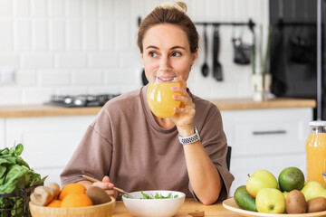 Wall Mural - healthy lifestyle. Happy young woman eating salad and drinking orange juice while sitting at home in the kitchen. Young female eat wholesome breakfast