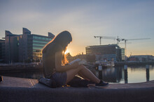 Young Girl With Long Hair Sitting On The Breakwater In The Harbor Reading A Book In The Light Of The Rising Sun, Golden Hour Photo Shoot, Dublin, Ireland