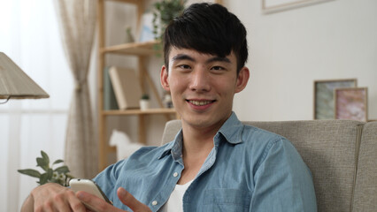 portrait of a smiling asian young male turning head to look at the camera while scrolling on his cell phone on the sofa in a modern living room at home.