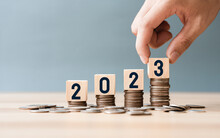 Hand Place Wooden Cube 2023 On Top Stack Of Coins With Sky Blue Background And Copy Space. Saving Money And Financial Plan Concept For Investment In New Year 2023.