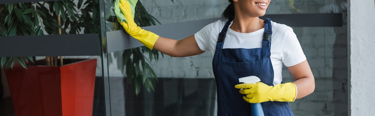 Wall Mural - cropped view of smiling bi-racial woman in rubber gloves cleaning glass door in office, banner.