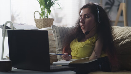 Woman with a disability in headphones sitting on a sofa writing in notebook and talking via video call while studying online on laptop at home