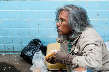 Wall Mural - An old, homeless, long-bearded Asian man sits on the street eating bread after receiving a delicious snack. from people passing by The poor man had no home in the streets.