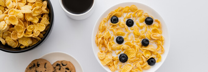 Wall Mural - top view of bowls with corn flakes in milk with blueberries near cup of coffee and cookies isolated on white, banner.