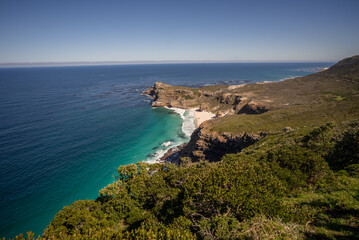  A wide shot over green vegetation to a stunning view of Dias beach, dramatic cliffs  and the blue sea from the western tip of Cape Point, Western Cape. 
