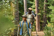 © pressmaster - Mature black man and his grandson with trekking sticks moving down footpath along green trees while enjoying backpack trip