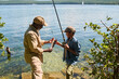 © pressmaster - Side view of happy mature black man in activewear showing fish to his grandson with rod while both standing against waterside