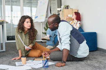 Aufkleber - Young confident businesswoman showing financial document to African American male coworker while both sitting on the floor in office