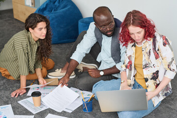 Aufkleber - Two intercultural economists looking at screen of laptop on knees of their colleague in casualwear sitting on the floor next to them