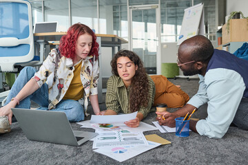 Aufkleber - Young African American man listening to female colleagues discussing financial documents while lying on office floor in front of laptop