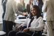 Portrait of a young African American businesswoman in a wheelchair smiling in the office - group of multi-ethnic businesspeople in the background - business lifestyle concept