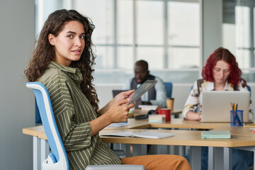 Poster - Young successful businesswoman with tablet sitting by workplace in office among her colleagues and looking at camera while networking