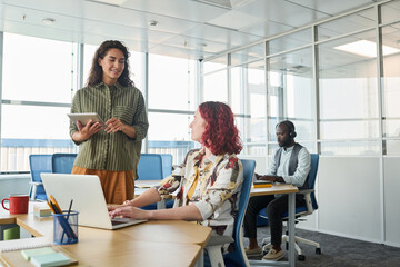 Wall Mural - Happy young office worker with digital tablet communicating with female colleague sitting by workplace in front of laptop during talk