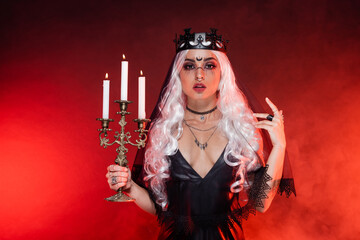 Wall Mural - blonde woman in black dress and witch crown holding triple candlestick on background with red smoke.