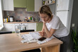 © DimaBerlin - Mature woman looks at communal payments bills placed on table. Thoughtful single female of middle age looks unsatisfied with utility bill sums. Blonde lady examines papers in kitchen closeup