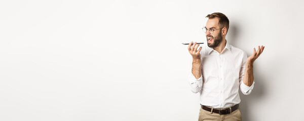 Wall Mural - Angry business man shouting at speakerphone, record voice message in mad state, standing against white background