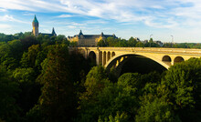 Scenic View Of Adolphe Bridge, Stone Arched Aqueduct Running Over Green Valley Of Petrusse River In Luxembourg City With State Savings Bank And Banking Museum In Background On Sunny Summer Day..
