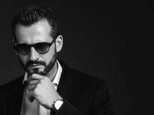 Closeup Portrait Of Handsome Smiling Stylish Hipster Model. Modern Man Dressed In Elegant Black Suit. Fashion Male Posing In Studio On Dark Background. In Spectacles. Black And White