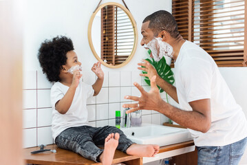 Wall Mural - Cheerful African American Father and little son laughing playing shaving foam on their faces in bathroom at home together, Black Family having fun.