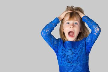 Wall Mural - Portrait of a little girl dressed in a blue dress on a gray background. The child poses for the camera with his hands on his head, depicting delight, surprise, joy.