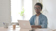 © stockbakers - Young African Man Looking at Camera while using Laptop in Office