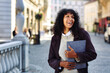 positive businesswoman standing on street holding folder with documents curly black hair