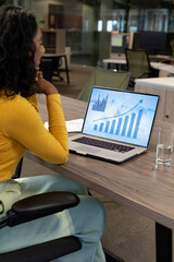 Sticker - Vertical of biracial businesswoman using laptop in empty office with graphs and data on screen