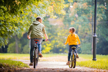 Happy father and daughter ride bicycles in autumn park on sunny day.