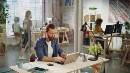 Poster - Handsome Long-Haired Bearded Manager Sitting at a Desk in Creative Office. Young Stylish Man Using Laptop Computer in Marketing Agency. Colleagues Working in the Background. High Angle View.