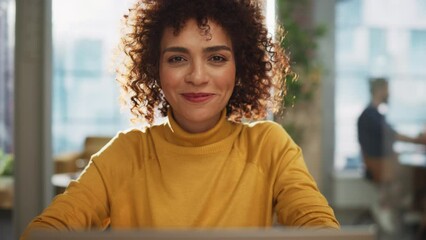 Wall Mural - Portrait of a Beautiful Middle Eastern Manager Sitting at a Desk in Creative Office. Young Stylish Female with Curly Hair Looking at Camera with Big Smile. Colleagues Working in the Background.