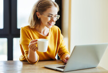 Cheerful Young Woman Uses A Laptop At Home In Kitchen With  Cup Of Morning Coffee