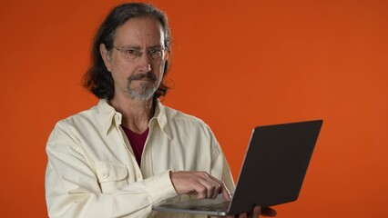 Portrait of smiling happy mature man on on laptop computer giving thumbs up with copy space on solid orange background.
