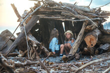 Two Little Cheerful Girls Sisters Of Different Ages Are Sitting In A Homemade Hut Made Of Logs And Sticks Of Trees On The Seashore