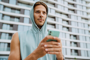 Wall Mural - Young athletic long-haired serious handsome man in hood with phone