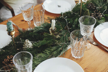 Stylish Christmas table setting. Rosemary branch on plate, vintage cutlery, wine glass, candles, fir branches with golden lights, pine cones on table. Holiday dinner, atmospheric table set