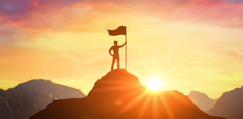 Silhouette of businessman holding flag on top mountain, sky and sun light background. Business success and goal concept. Business man with victory flag on hilltop at sunset