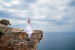 © svetograph - Blonde with long hair on a sunny seashore in a white flowing dress, rear view, silk fabric waving in the wind. Against the backdrop of the blue sky and mountains on the seashore.